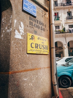 The image features a street corner with a textured wall and several signs. There is a vibrant blue street sign in Arabic and French, along with a vintage yellow advertisement for an orange juice drink called 'CRUSH'. A section of a white building with balconies is visible in the background, accompanied by parked cars including a blue vehicle.