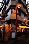 A street scene featuring a Japanese building corner adorned with traditional artwork. Signs with Japanese characters and colorful lanterns are visible, casting a warm glow. Several advertisements and plaques are attached to the building, and people are seated inside a lit-up restaurant visible through the windows.