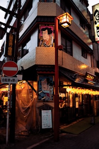 A street scene featuring a Japanese building corner adorned with traditional artwork. Signs with Japanese characters and colorful lanterns are visible, casting a warm glow. Several advertisements and plaques are attached to the building, and people are seated inside a lit-up restaurant visible through the windows.