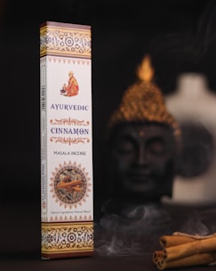 A box of Ayurvedic cinnamon masala incense is displayed prominently against a dark background. The packaging is elaborate with intricate patterns and warm colors, featuring an image of a person in traditional attire. To the side, there is a Buddha statue slightly out of focus, creating a serene setting. Cinnamon sticks and gentle wafts of incense smoke add to the ambiance.