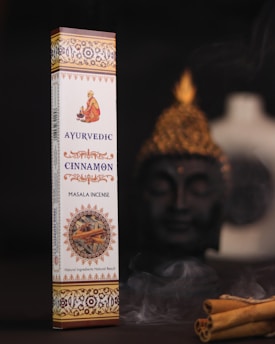 A box of Ayurvedic cinnamon masala incense is displayed prominently against a dark background. The packaging is elaborate with intricate patterns and warm colors, featuring an image of a person in traditional attire. To the side, there is a Buddha statue slightly out of focus, creating a serene setting. Cinnamon sticks and gentle wafts of incense smoke add to the ambiance.