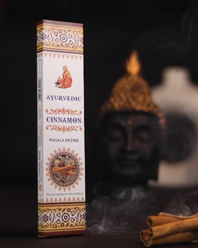 A box of Ayurvedic cinnamon masala incense is displayed prominently against a dark background. The packaging is elaborate with intricate patterns and warm colors, featuring an image of a person in traditional attire. To the side, there is a Buddha statue slightly out of focus, creating a serene setting. Cinnamon sticks and gentle wafts of incense smoke add to the ambiance.