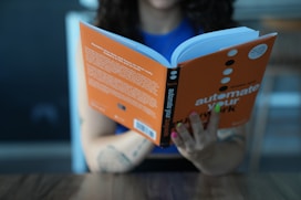 A person is reading a book with an orange cover titled 'automate your work'. The person's fingernails are painted various colors, and they are seated at a table with a wooden surface. The background appears to be blurred, possibly indicating an indoor setting.