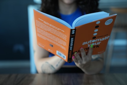 A person is reading a book with an orange cover titled 'automate your work'. The person's fingernails are painted various colors, and they are seated at a table with a wooden surface. The background appears to be blurred, possibly indicating an indoor setting.