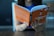 A person is reading a book with an orange cover titled 'automate your work'. The person's fingernails are painted various colors, and they are seated at a table with a wooden surface. The background appears to be blurred, possibly indicating an indoor setting.