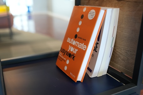 A close-up view of a stack of books on a dark shelf, with the most prominent book having an orange cover and white bold text reading 'automate your busywork'. The bookshelf is modern and minimalist in style, placed against a wooden backdrop.