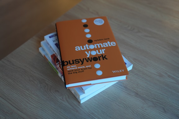 A stack of books on a wooden surface, with the top book displaying a bold, orange cover featuring the title 'automate your busywork' in large white and black letters.