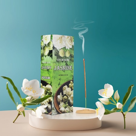 A box of jasmine incense sticks stands vertically on a round platform. The box is green with images of jasmine flowers and includes text indicating its Ayurvedic qualities. An incense stick is lit and releases a thin trail of smoke. White jasmine flowers are placed decoratively around the box, adding a serene touch. The background is a calming teal color.