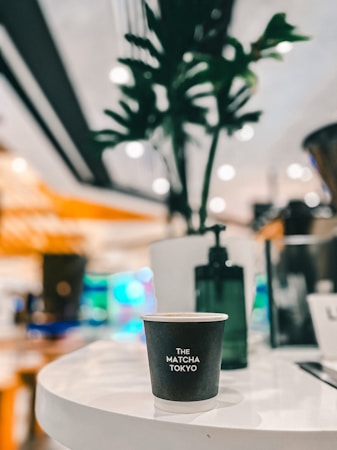 A takeaway coffee cup with the text 'The Matcha Tokyo' is placed on a glossy white table. There is a blurred plant in the background, and the environment seems to be a busy, well-lit cafe or restaurant setting.