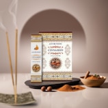Boxes of Ayurvedic cinnamon masala incense are artfully displayed with intricate patterns and a central image of cinnamon sticks. Nearby, incense sticks emit gentle smoke, while bowls of ground cinnamon and loose sticks are arranged alongside nutmeg and star anise, set against a soft, neutral background.
