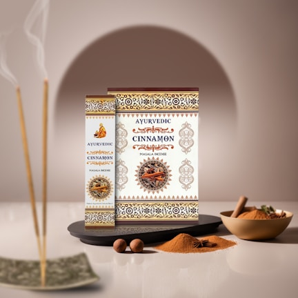 Boxes of Ayurvedic cinnamon masala incense are artfully displayed with intricate patterns and a central image of cinnamon sticks. Nearby, incense sticks emit gentle smoke, while bowls of ground cinnamon and loose sticks are arranged alongside nutmeg and star anise, set against a soft, neutral background.