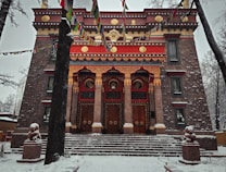 A large, ornately decorated building with traditional architecture featuring intricate patterns and vivid colors. The structure is flanked by two trees and adorned with colorful prayer flags. Snow covers the ground and the branches of the trees, creating a serene winter scene.