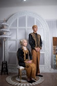 A man and a woman dressed in traditional attire pose in a formal setting. The woman is seated on a chair, while the man stands beside her. The woman wears a black outfit with gold patterns and a headscarf, and the man wears a matching black and gold outfit. They are in front of a decorative white and gold backdrop.