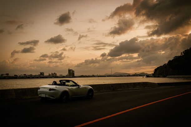A convertible sports car is parked by the roadside facing a large body of water. The sky is filled with dramatic, thick clouds, and the lighting suggests either sunrise or sunset. In the distance, an industrial skyline is visible along the horizon, with a bridge extending across part of the water.
