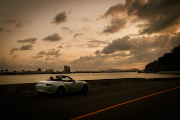 A convertible sports car is parked by the roadside facing a large body of water. The sky is filled with dramatic, thick clouds, and the lighting suggests either sunrise or sunset. In the distance, an industrial skyline is visible along the horizon, with a bridge extending across part of the water.