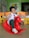 A young child wearing a school uniform is sitting on a red rocking horse toy in a colorful classroom environment. The child is smiling playfully and gesturing with their hands. Surrounding the child are bright yellow and orange chairs, and the walls are decorated with cartoon characters.
