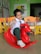 A young child wearing a school uniform is sitting on a red rocking horse toy in a colorful classroom environment. The child is smiling playfully and gesturing with their hands. Surrounding the child are bright yellow and orange chairs, and the walls are decorated with cartoon characters.
