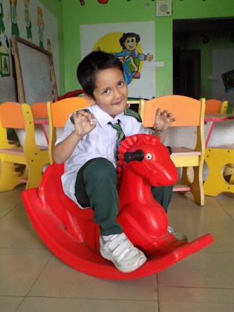 A young child wearing a school uniform is sitting on a red rocking horse toy in a colorful classroom environment. The child is smiling playfully and gesturing with their hands. Surrounding the child are bright yellow and orange chairs, and the walls are decorated with cartoon characters.