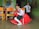 Two young children in school uniforms are indoors. One child is seated on a red rocking horse, wearing a pink backpack, and appears cheerful. The other child stands behind, smiling, near colorful furniture. The walls have a mural with playful illustrations.
