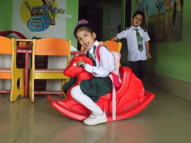 Two young children in school uniforms are indoors. One child is seated on a red rocking horse, wearing a pink backpack, and appears cheerful. The other child stands behind, smiling, near colorful furniture. The walls have a mural with playful illustrations.