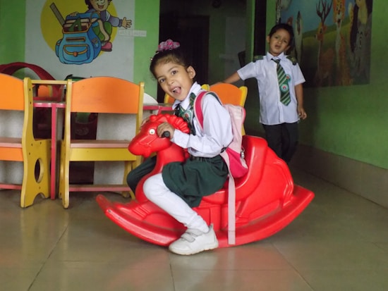 Two young children in school uniforms are indoors. One child is seated on a red rocking horse, wearing a pink backpack, and appears cheerful. The other child stands behind, smiling, near colorful furniture. The walls have a mural with playful illustrations.