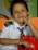 A young child wearing a school uniform with a green-striped tie and a shirt marked 'KG' sits in an indoor environment with colorful backgrounds. The child has a playful expression, with one hand resting on a bright red toy.