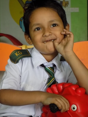 A young child wearing a school uniform with a green-striped tie and a shirt marked 'KG' sits in an indoor environment with colorful backgrounds. The child has a playful expression, with one hand resting on a bright red toy.