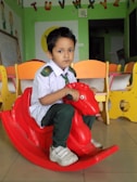 A young child is sitting on a red plastic rocking toy shaped like a horse in a classroom. The child is wearing a school uniform, consisting of a white shirt, dark green pants, a green tie, and white sneakers. In the background, there are colorful child-sized chairs and educational decorations, including the alphabet letters attached to the wall.