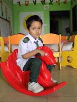 A young child is sitting on a red plastic rocking toy shaped like a horse in a classroom. The child is wearing a school uniform, consisting of a white shirt, dark green pants, a green tie, and white sneakers. In the background, there are colorful child-sized chairs and educational decorations, including the alphabet letters attached to the wall.