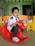 A young child is sitting on a red plastic rocking toy shaped like a horse in a classroom. The child is wearing a school uniform, consisting of a white shirt, dark green pants, a green tie, and white sneakers. In the background, there are colorful child-sized chairs and educational decorations, including the alphabet letters attached to the wall.