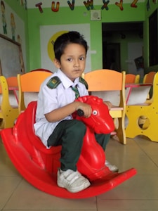 A young child is sitting on a red plastic rocking toy shaped like a horse in a classroom. The child is wearing a school uniform, consisting of a white shirt, dark green pants, a green tie, and white sneakers. In the background, there are colorful child-sized chairs and educational decorations, including the alphabet letters attached to the wall.