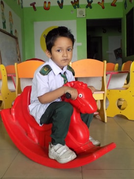 A young child is sitting on a red plastic rocking toy shaped like a horse in a classroom. The child is wearing a school uniform, consisting of a white shirt, dark green pants, a green tie, and white sneakers. In the background, there are colorful child-sized chairs and educational decorations, including the alphabet letters attached to the wall.