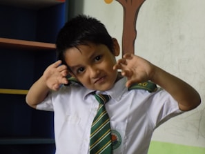 A young child dressed in a school uniform poses playfully with hands near the face. The uniform includes a white shirt and a green striped tie, with initials 'KG' visible on the tie. The child has short dark hair and stands in a room with a partially visible bookshelf and a wall that has a tree drawing.