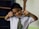 A young child dressed in a school uniform poses playfully with hands near the face. The uniform includes a white shirt and a green striped tie, with initials 'KG' visible on the tie. The child has short dark hair and stands in a room with a partially visible bookshelf and a wall that has a tree drawing.
