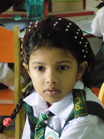 A young girl with braided hair adorned with a band featuring small decorative beads is sitting indoors. She is wearing a school uniform consisting of a white shirt and a green tie with yellow stripes, with a patch on her shirt indicating a school name. The background shows a classroom setting with colorful furniture.