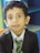 A young child with short dark hair is wearing a black suit jacket, white dress shirt, and a green tie with yellow stripes. The background features a multicolored environment with blue and green elements.