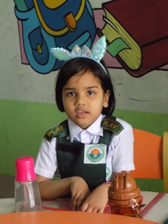 A young girl in a school uniform is sitting at a table. She is wearing a headband with sparkling decorative pieces. On the table, there is a pink water bottle and a brown container. The background has colorful illustrations of a backpack and books.