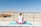 A person is seated on a blue yoga mat in a meditative pose, facing a vast desert landscape. The person is wearing a sleeveless white top and pink pants, with hair tied up. In the distance, there are rugged mountains and a clear blue sky. To the left, there are structures resembling rustic cabins made of wood.
