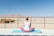 A person is seated on a blue yoga mat in a meditative pose, facing a vast desert landscape. The person is wearing a sleeveless white top and pink pants, with hair tied up. In the distance, there are rugged mountains and a clear blue sky. To the left, there are structures resembling rustic cabins made of wood.