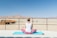 A person is seated on a blue yoga mat in a meditative pose, facing a vast desert landscape. The person is wearing a sleeveless white top and pink pants, with hair tied up. In the distance, there are rugged mountains and a clear blue sky. To the left, there are structures resembling rustic cabins made of wood.