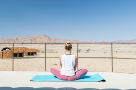 A person is seated on a blue yoga mat in a meditative pose, facing a vast desert landscape. The person is wearing a sleeveless white top and pink pants, with hair tied up. In the distance, there are rugged mountains and a clear blue sky. To the left, there are structures resembling rustic cabins made of wood.
