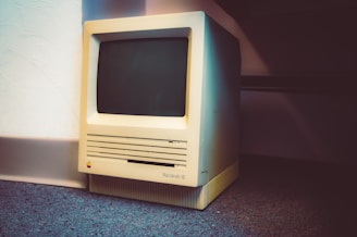 A vintage Macintosh SE computer is placed on a carpeted floor against a white wall. The old computer features a small screen and a beige casing, with visible vents and a multicolored Apple logo.