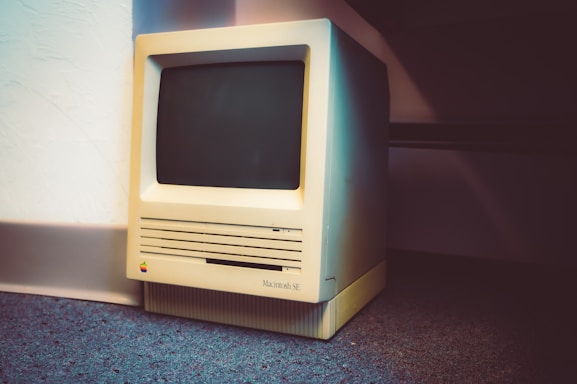 A vintage Macintosh SE computer is placed on a carpeted floor against a white wall. The old computer features a small screen and a beige casing, with visible vents and a multicolored Apple logo.