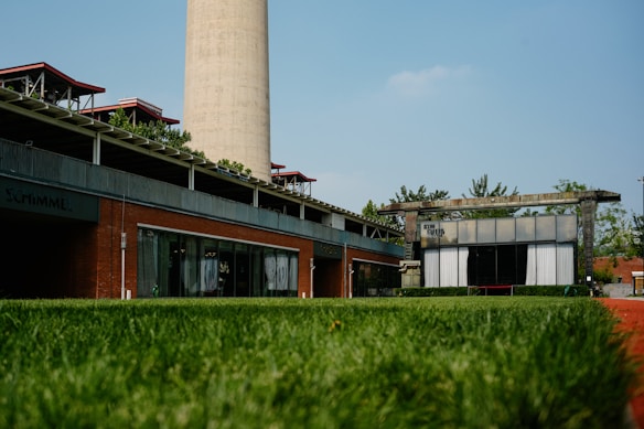 An industrial building with modern architecture featuring brick and steel elements. A large smokestack rises in the background, while a lush green lawn dominates the foreground. Trees and small shrubs contribute to the greenery surrounding the area.