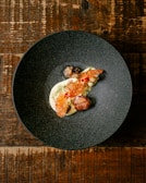 A dark, textured bowl contains a gourmet dish with artistic presentation. The dish includes a creamy white base, topped with a red, lattice-like garnish. There is a round, browned element resembling a meatball and small vegetable components. The bowl rests on a rustic wooden table.