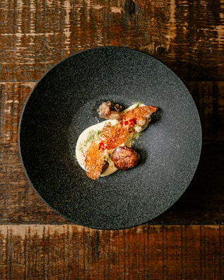 A dark, textured bowl contains a gourmet dish with artistic presentation. The dish includes a creamy white base, topped with a red, lattice-like garnish. There is a round, browned element resembling a meatball and small vegetable components. The bowl rests on a rustic wooden table.
