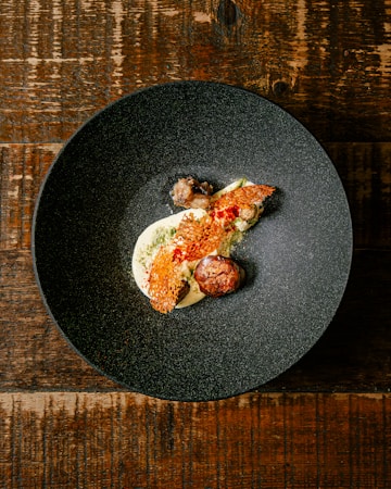 A dark, textured bowl contains a gourmet dish with artistic presentation. The dish includes a creamy white base, topped with a red, lattice-like garnish. There is a round, browned element resembling a meatball and small vegetable components. The bowl rests on a rustic wooden table.