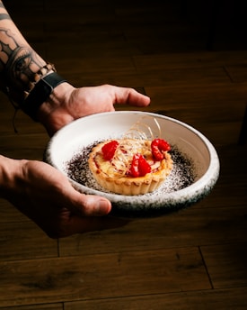 A person is holding a white ceramic plate with a beautifully presented tart. The tart is topped with cream, fresh raspberries, and decorative spun sugar. The plate has a sprinkle of dark chocolate crumbs around the edges, adding contrast. The background is a wooden floor, providing a warm and rustic feel.