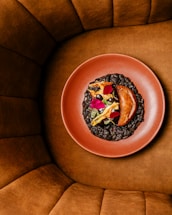A aesthetically plated dish in a red clay bowl, featuring a piece of cooked meat accompanied by gourmet garnishes such as flower petals and vegetables. The bowl is surrounded by a plush, brown cushioned seating area.
