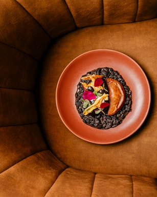 A aesthetically plated dish in a red clay bowl, featuring a piece of cooked meat accompanied by gourmet garnishes such as flower petals and vegetables. The bowl is surrounded by a plush, brown cushioned seating area.